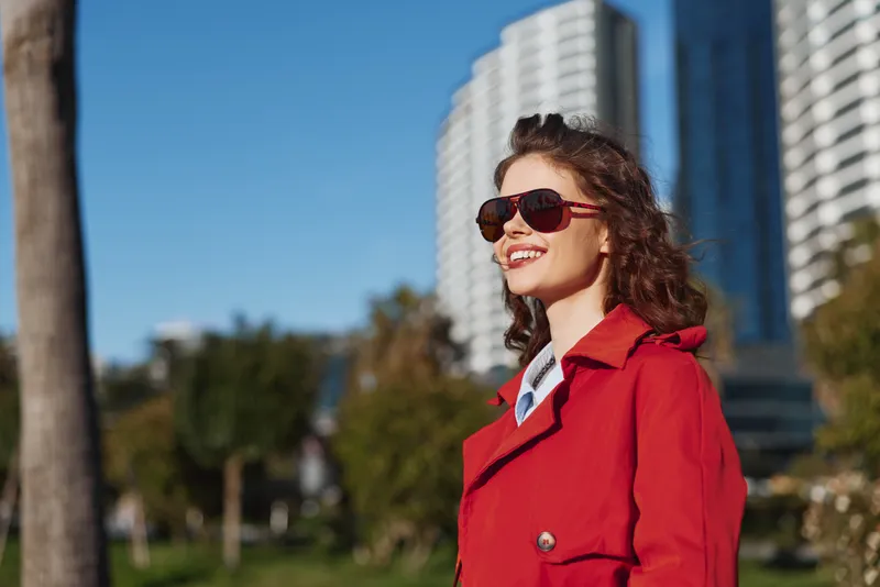 Young woman with curly hair smiles confidently wearing a red coat and sunglasses outdoors on a sunny day with modern buildings in the background.