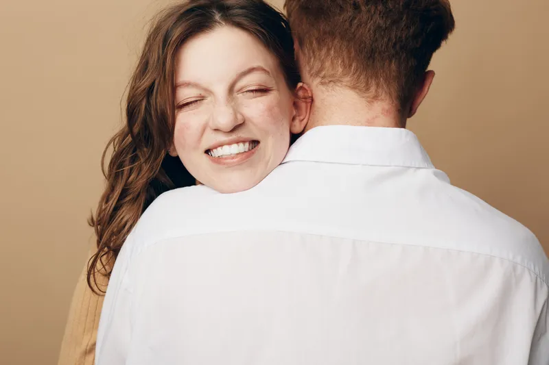 Happy woman with closed eyes smiles while hugging man in white shirt. Intimate moment of affection and love between couple on beige background. Concept of relationship and emotional connection.