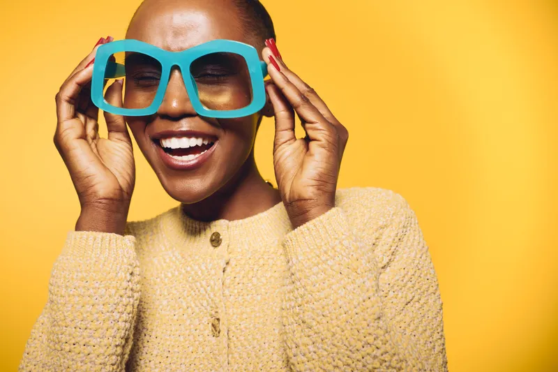 Happy African woman with dark skin wearing oversized turquoise glasses and beige sweater smiles joyfully against yellow background. Concept of fashion, style, and positive emotions.