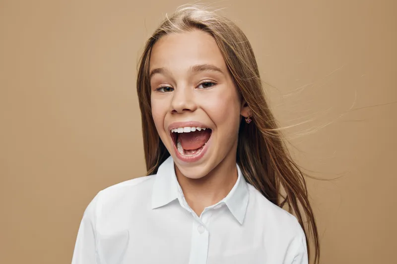 Girl with long hair smiling and laughing against beige background wearing white shirt in casual portrait showing happy emotions and open mouth expression.