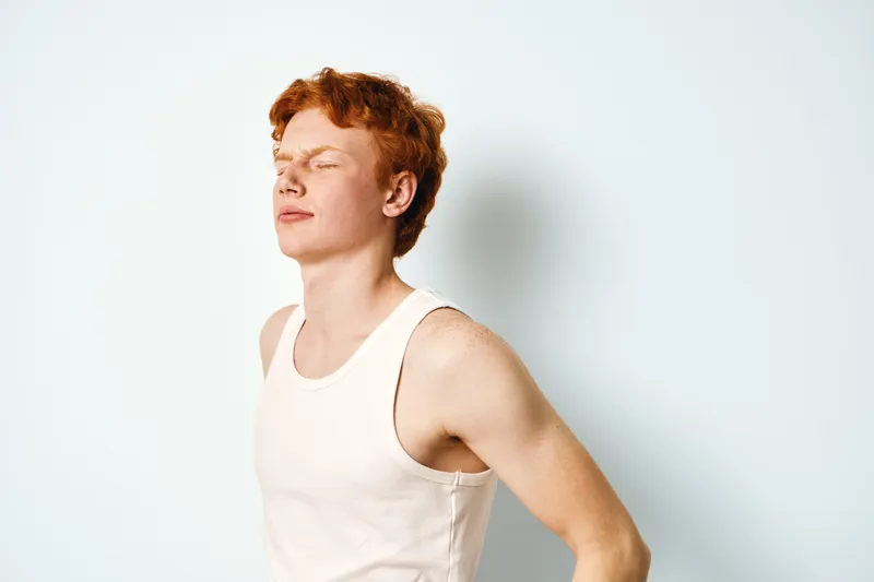 Young man with red hair and freckles wearing white tank top standing against white background with closed eyes and relaxed expression. Portrait of casual style person.