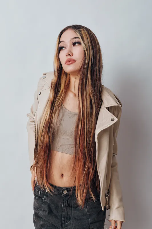 Young woman with long hair wearing beige jacket and crop top posing against white background. Casual style, fashion, confident, standing with hands in pockets and looking away.