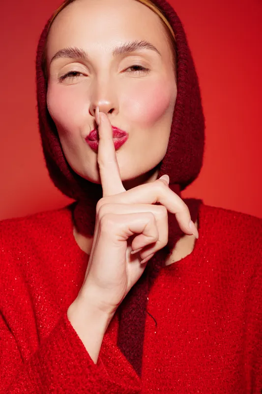 Woman in red sweater and hood makes silence gesture with finger on lips against red background. Concept of secrecy, quiet, and calm expression in studio portrait.