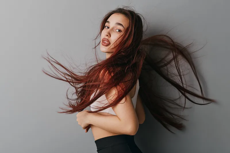 Woman with long flowing hair posing against gray background, fashion model in casual outfit with dynamic movement and natural makeup, studio portrait with soft lighting.