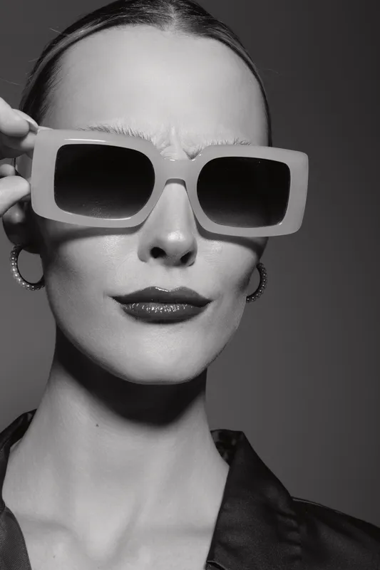 Close-up portrait of a pale Caucasian woman wearing large sunglasses and hoop earrings. Stylish fashion concept with confident expression in black and white studio lighting.