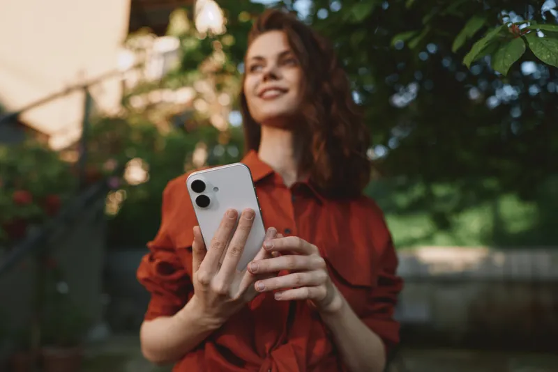 A woman outdoors holding a smartphone, smiling in a casual outfit. A portrait style lifestyle scene emphasizing joy, connection, and everyday technology