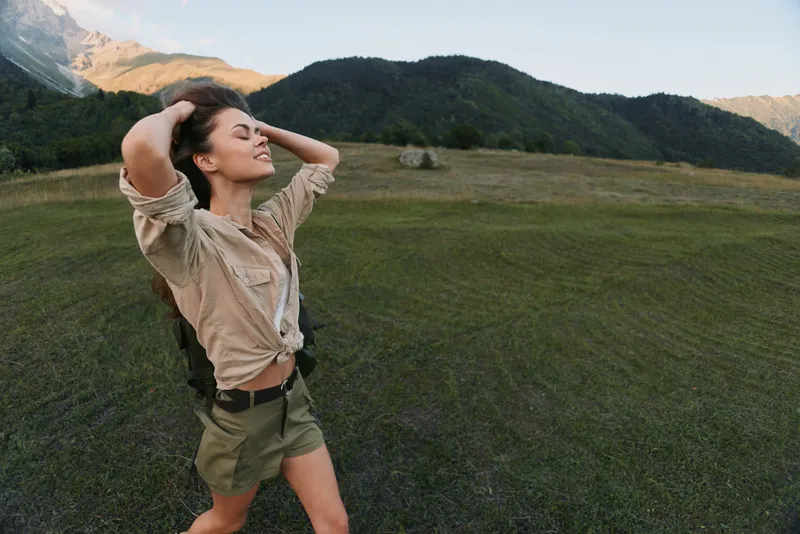Young woman with long hair enjoys a carefree moment outdoors in casual shorts and a tied shirt with a backpack in a mountain meadow during a sunny day.