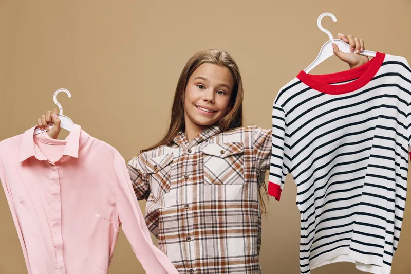 Girl smiling holding two hangers with pink shirt and striped sweater for clothing choice, fashion, shopping and style on beige background. Young female with casual outfit.