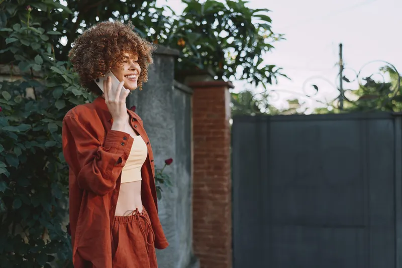 A woman outdoors wears a red casual outfit, smiling while talking on a phone in a sunlit yard. A lifestyle moment capturing everyday communication, confidence, and modern style