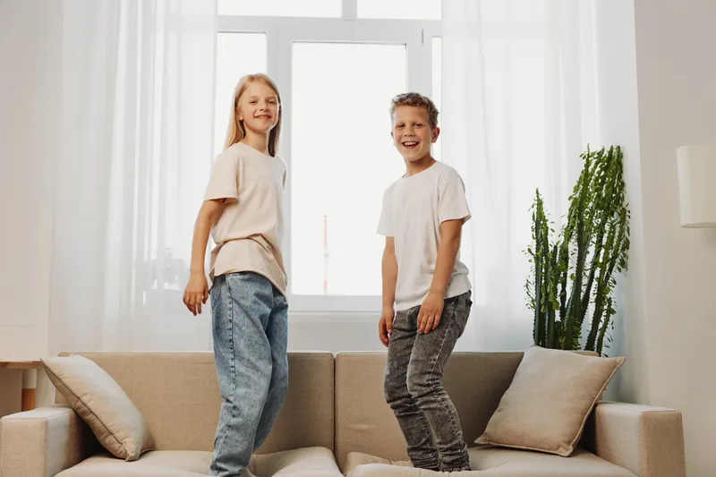 Two cheerful children jumping on a beige sofa in a bright living room with natural light and green plant decor.