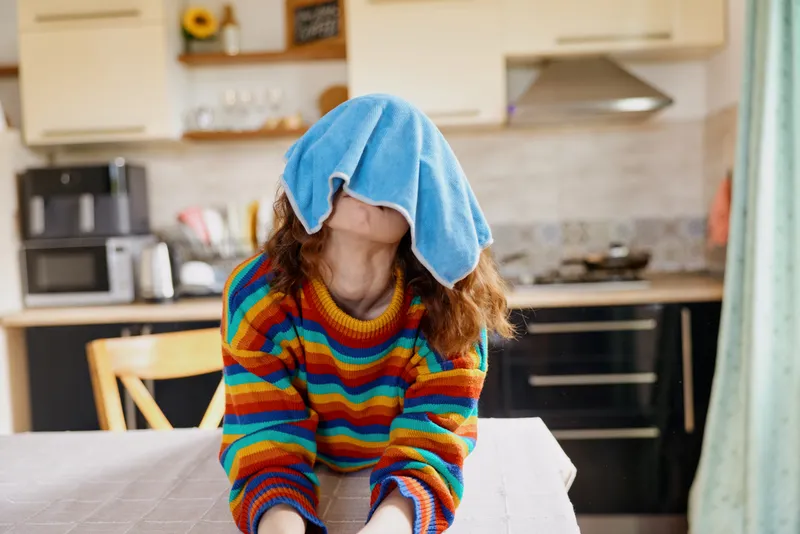 Playful girl with a towel over her head sits at a kitchen table, wearing a bright striped sweater, creating a lighthearted home moment and playful family scene.