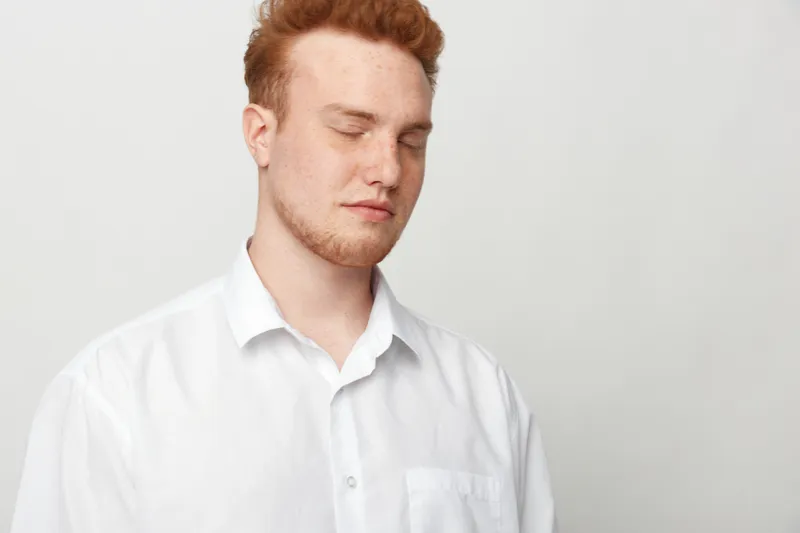 Redhead man with closed eyes wearing white shirt stands against plain light background. Calm expression and relaxed posture create peaceful and introspective mood in studio portrait.