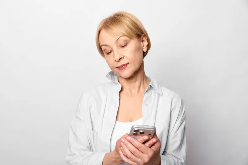 Senior woman with smartphone indoors, checking messages on screen, gentle smile, relaxed posture, soft studio light, neutral background, candid portrait