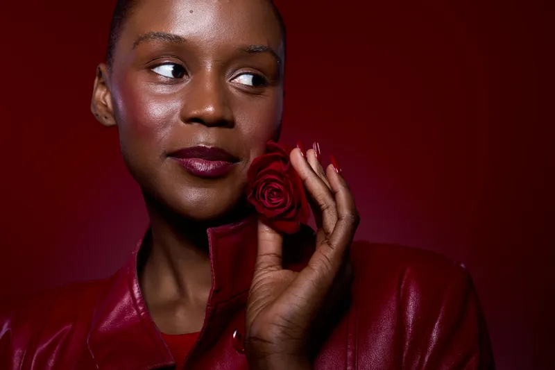 Close-up of a confident dark skinned African woman wearing a red leather jacket holding a red rose near her face. Concept of beauty, elegance, and self-confidence with red background.