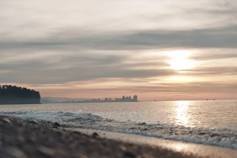 Beach at sunset with calm water and gentle waves, distant city skyline on the horizon, warm light reflecting on wet sand, serene coastal scene suitable for travel and nature visuals
