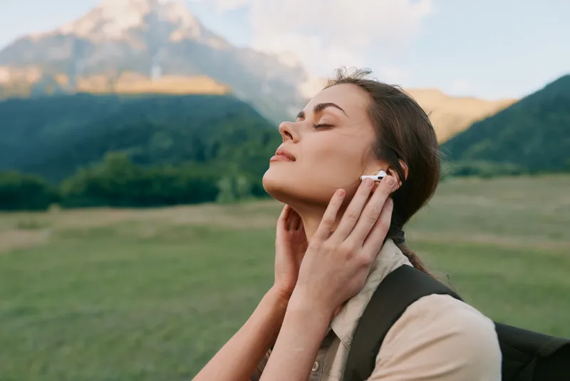 Young woman outdoors in a grassy field, eyes closed, listening to headphones, as distant mountains glow in the morning light, conveying calm, focus, and personal wellness.