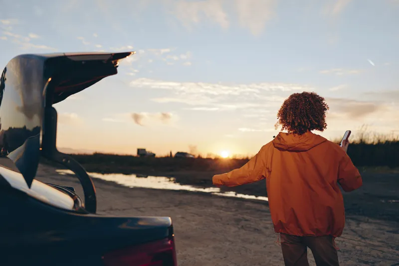 Woman lifestyle outdoors sunset, standing by car, travel moment, holding phone, walking along road, warm golden light, relaxed casual jacket, soft breeze, open sky, distant fields, calm cinematic mood