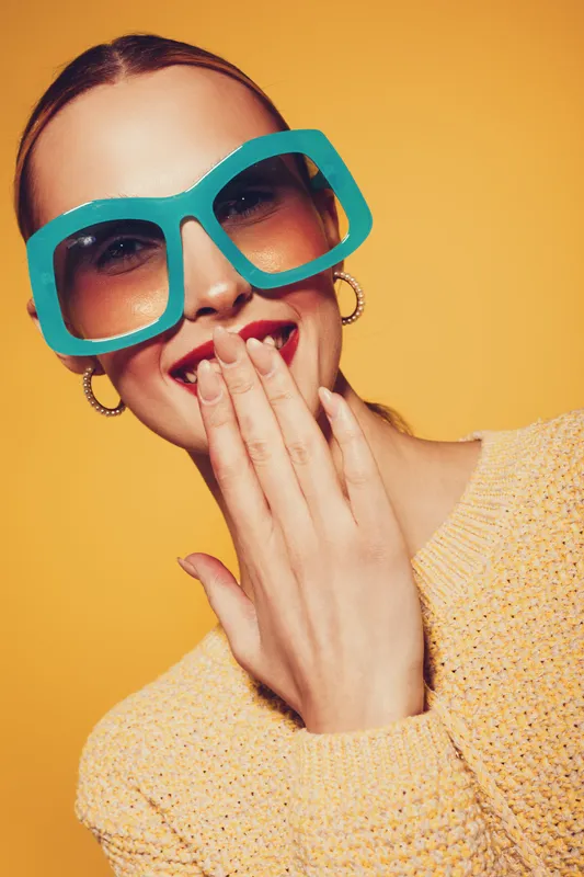Young woman with large turquoise sunglasses laughs covering mouth with hand. She wears hoop earrings and a yellow sweater against a bright yellow background. Concept of joy and fashion.