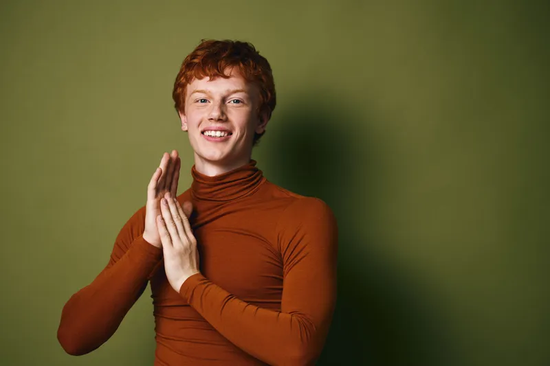 Smiling young man with red hair wearing brown turtleneck clapping hands in front of green background with happy expression for casual portrait and positive emotion concept.