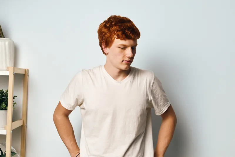 Young man with red hair wearing white t shirt standing with hands on hips looking down in bright studio with light background and decorative shelves.