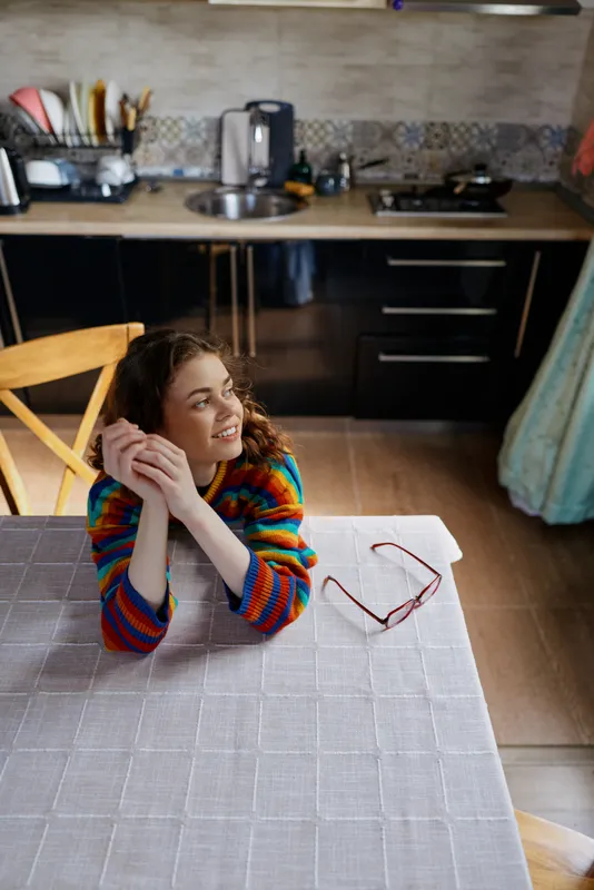 A woman rests her chin on her hands at a table in a bright kitchen, wearing a colorful striped sweater, glasses on the table, a cozy home moment of relaxation and simple living
