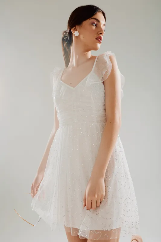 Young woman in a white lace dress poses gracefully looking to the side. Soft lighting highlights her beauty and fashion style in a minimal studio setting.