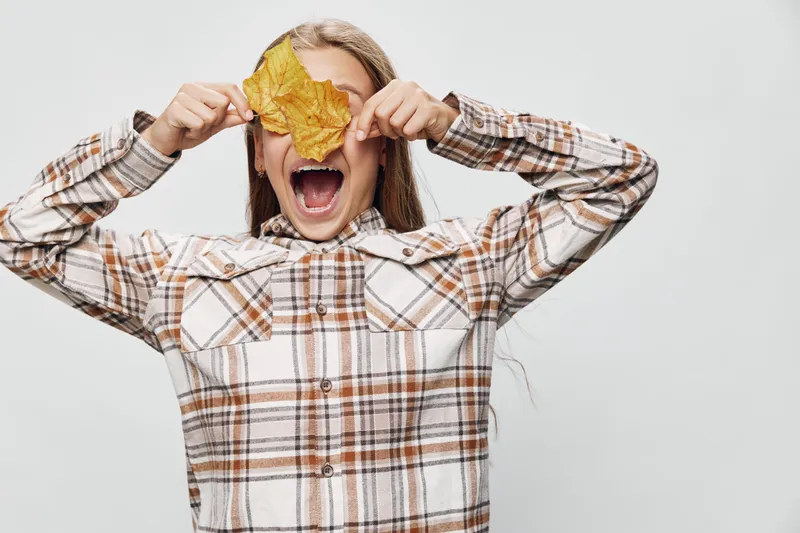 Girl in checkered shirt holding yellow autumn leaf covering eye with open mouth on white background. Child playing, happy with seasonal nature and fall mood for activity outdoors.