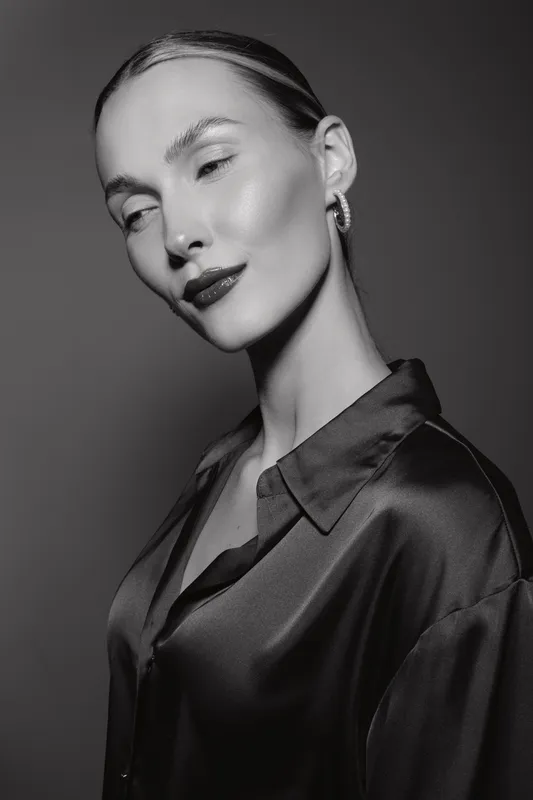 Portrait of a young Caucasian woman with fair skin wearing a silk blouse and hoop earrings on a dark background.