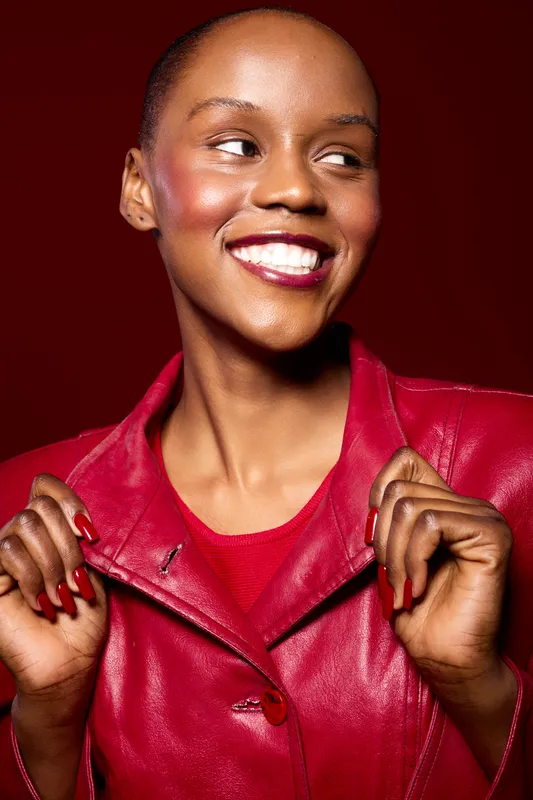 Smiling African woman with dark skin wearing a red leather jacket looks away confidently. Studio portrait with bright makeup and glossy lips on deep red background. Fashion and beauty.