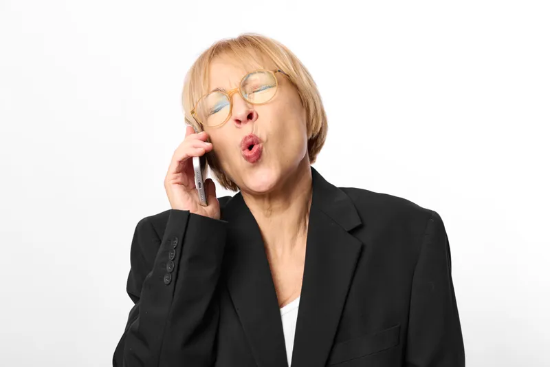 Older woman with glasses in black blazer, talking on phone, surprised expression, studio headshot against white background, business communication concept