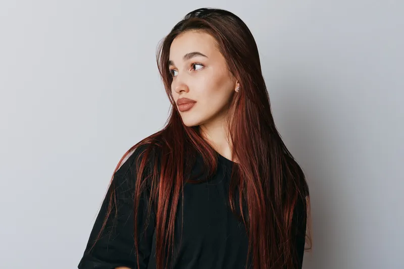 Young woman with long red hair wearing black shirt standing against white background looking sideways with thoughtful expression and natural makeup in studio portrait.