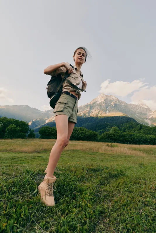 Young woman with a backpack leaps across a grassy field as mountains rise behind, capturing a lively outdoor hiking moment under pale blue sky with soft light and a gentle breeze gently.