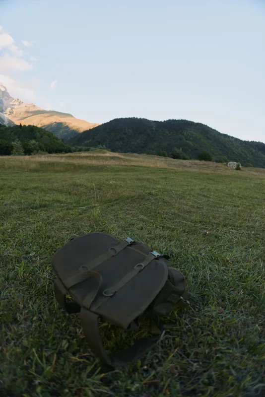 A lone backpack lies on a grassy meadow with distant hills and a clear sky, conveying outdoor travel, exploration, and a sense of nature escape.