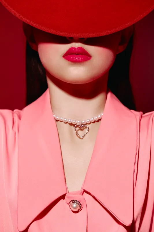 Stylish woman wearing a vibrant red hat and pink blazer, showcasing bold pink lips and a pearl necklace with a heart pendant. Concept for fashion, beauty, and elegance.