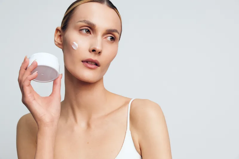 Young woman with clear skin holding a white jar of face cream applying moisturizer on cheek. Studio portrait on light background. Concept of skincare beauty wellness and self-care.