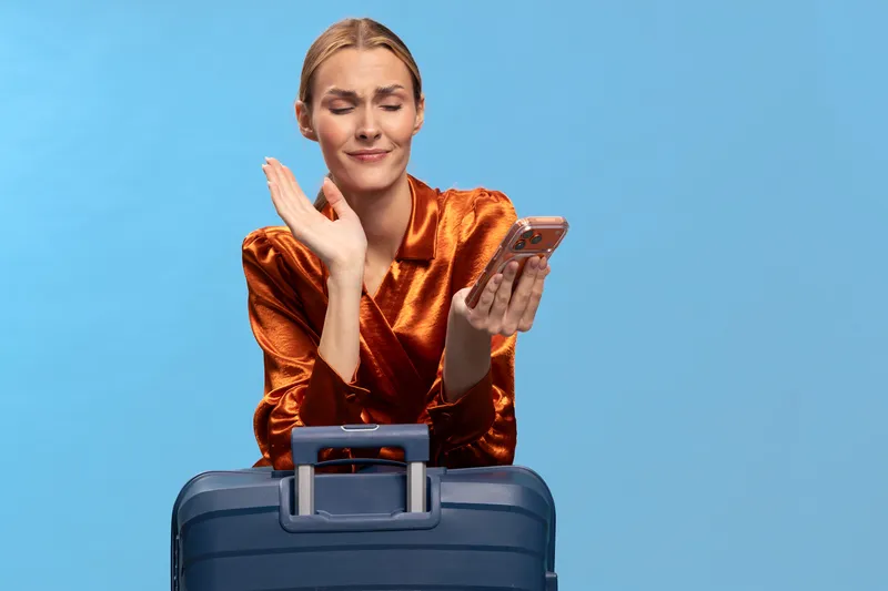Frustrated woman in orange satin blouse holds smartphone while standing behind blue suitcase against blue background. Concept of travel delays, communication problems, and stress during trip.