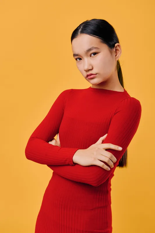 Young woman with long dark hair in red dress stands with crossed arms against orange background. Serious confident look, minimalistic style, portrait of thoughtful female model in studio.