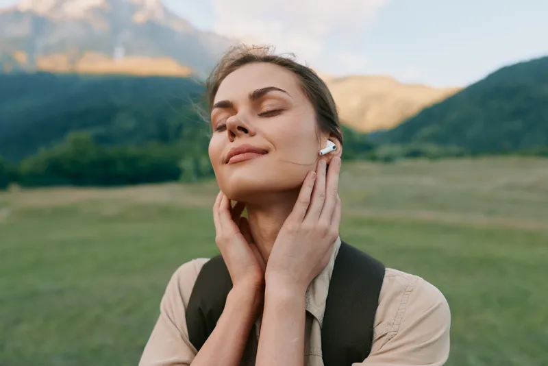 Woman enjoying outdoor life with earbud, eyes closed, serene mood, standing in a meadow near mountains, showcasing a calm nature lifestyle moment