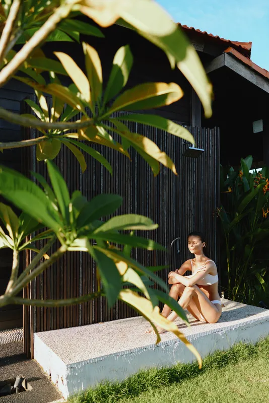 Young woman sits on concrete steps beside a wooden house, surrounded by tropical plants, enjoying a warm sunlit outdoor moment with a relaxed, serene mood and gentle breeze