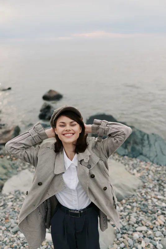 Smiling woman in checkered coat and white shirt stands on rocky beach with sea in background. Relaxed mood, casual fashion, outdoor lifestyle, natural light, happy female portrait.