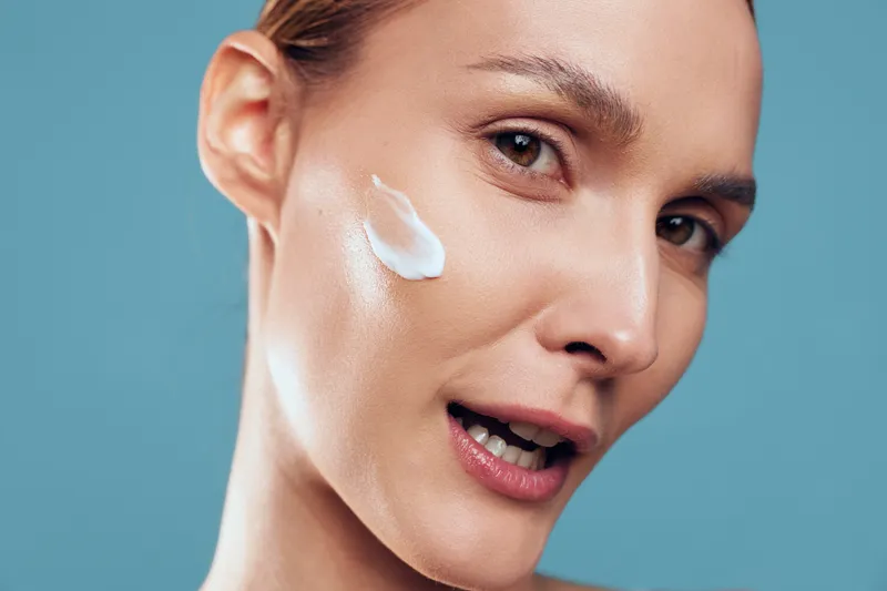 Close-up of a young woman applying white face cream on her cheek against a blue background. Concept of skincare, beauty, wellness, and self-care with natural lighting.