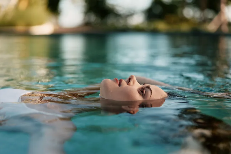 Tranquil woman floats on her back in a clear pool, sunlit water reflects greenery, capturing a relaxed lifestyle moment full of calm and bliss and gentle warmth