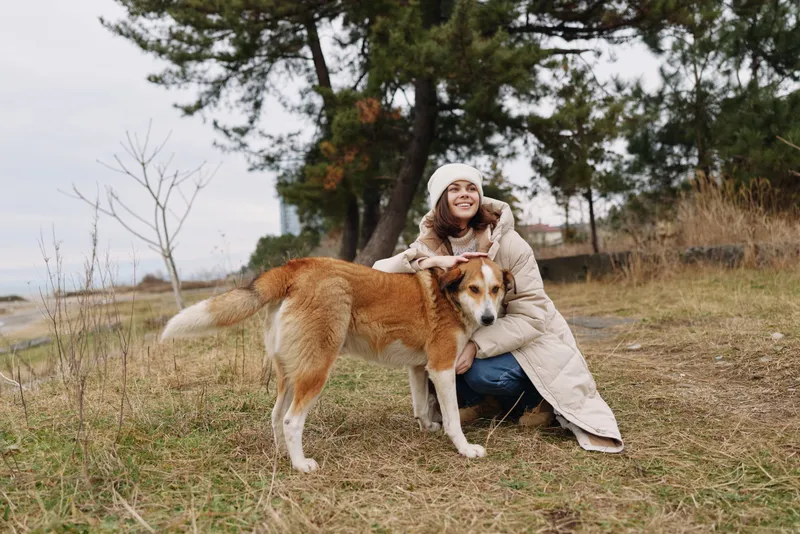 Woman kneeling beside a large dog in an autumnal park, wearing a warm coat and hat, smiling as they bond in an open field with trees and overcast sky together.