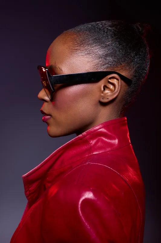 Profile of a confident dark skinned African woman wearing large sunglasses and a red leather jacket against a dark background. Concept of fashion, style, and attitude.