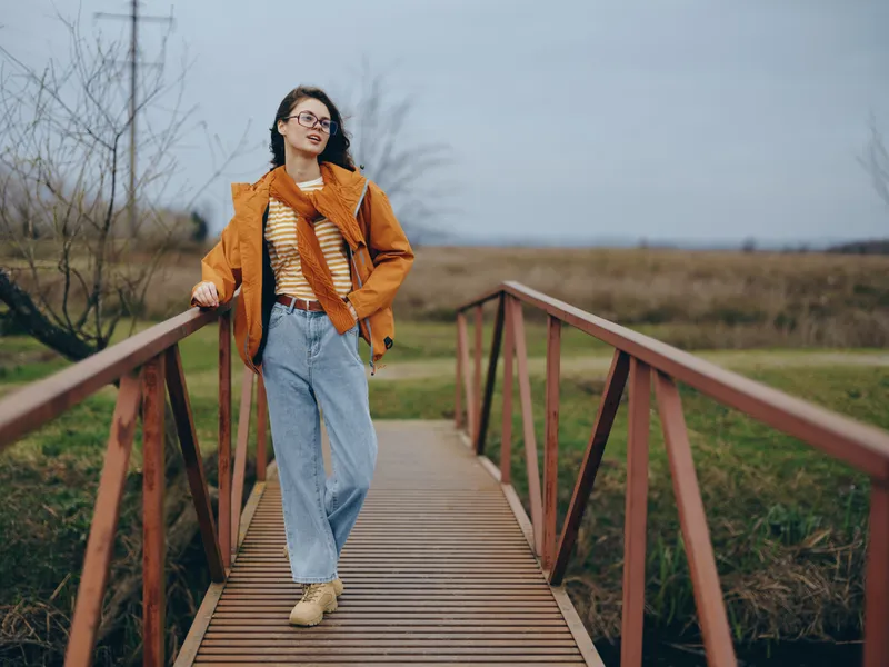 Woman lifestyle outdoors, walking on wooden boardwalk, orange jacket and scarf, casual jeans, smiling, glasses, soft cloudy light, spring field, relaxed posture, natural colors, today