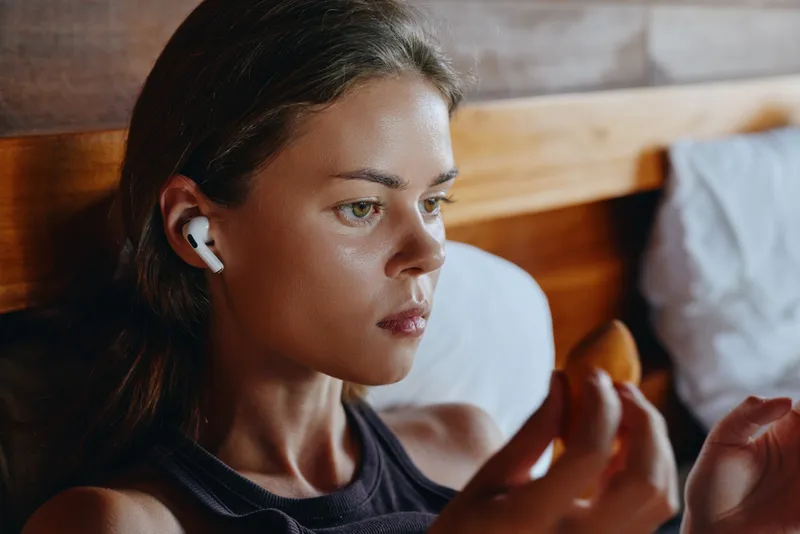 Calm woman wearing wireless earbuds sits on a bed in a softly lit room, focused on her device, creating a relaxed lifestyle moment with intimate personal tech use