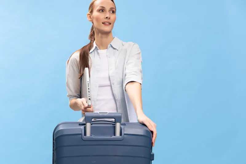 Young woman with long hair holding a laptop and a large suitcase stands against blue background. Concept of travel, business trip, and modern lifestyle with casual clothing and confidence.