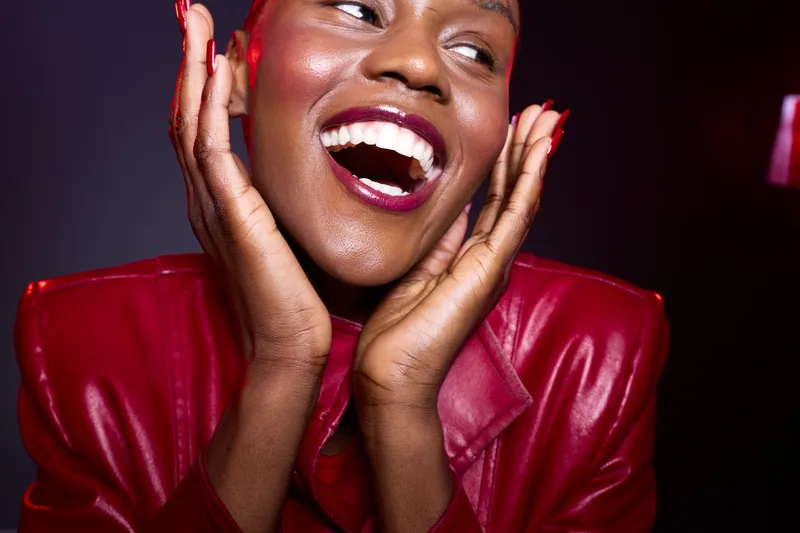 Close-up portrait of a happy black woman with dark skin wearing a red leather jacket smiling joyfully with hands near face on dark background. Concept of happiness, fashion, and beauty.