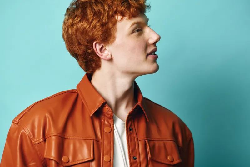 Young man with red hair wearing brown leather jacket and white shirt looking sideways on blue background. Profile portrait with casual style and bright natural color contrast.