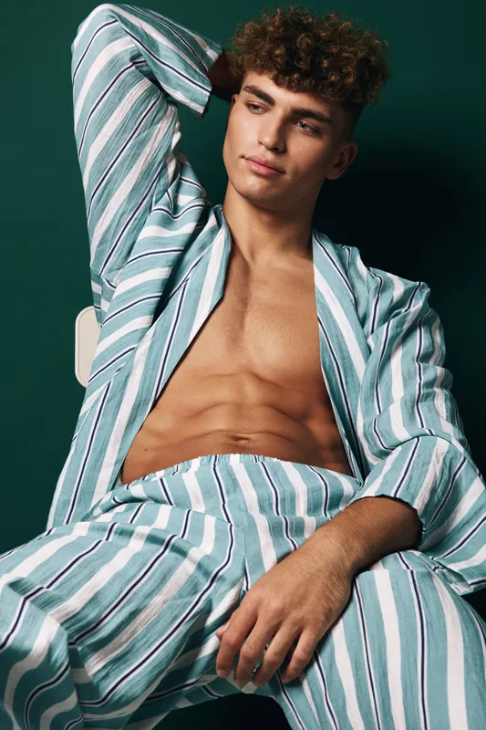 Young man with curly hair wearing striped pajamas sitting relaxed with hand behind head against dark background. Casual style and comfortable homewear.